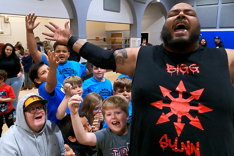 Big Synn strikes a pose with arms outstretched, surrounded by an excited crowd of young fans at a PCW event in the Oroville Convention Center.