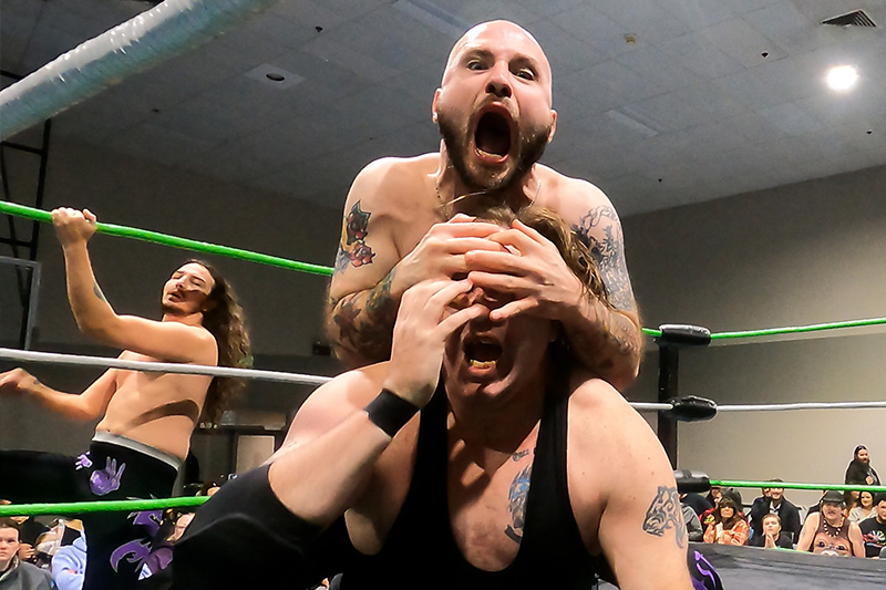 Nasty Nash of the Unwanted rakes Phil's face with his hands while shouting into the camera, with teammate Christian Skye observing from the ring apron at a PCW event in Red Bluff, CA.